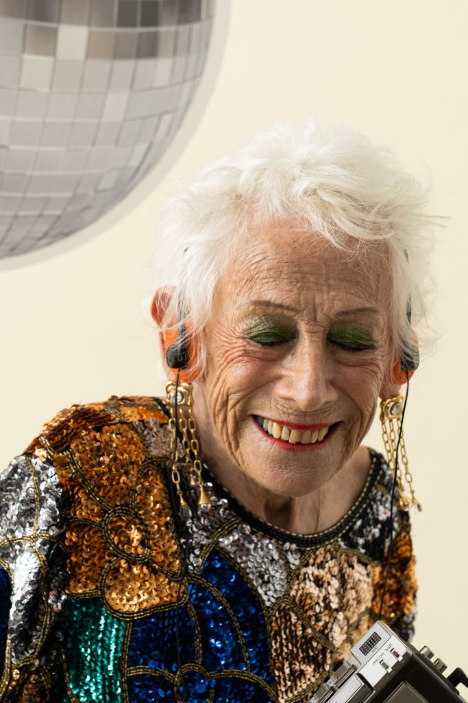 An older woman in a sequinned top smiling with her eyes closed beneath a disco ball