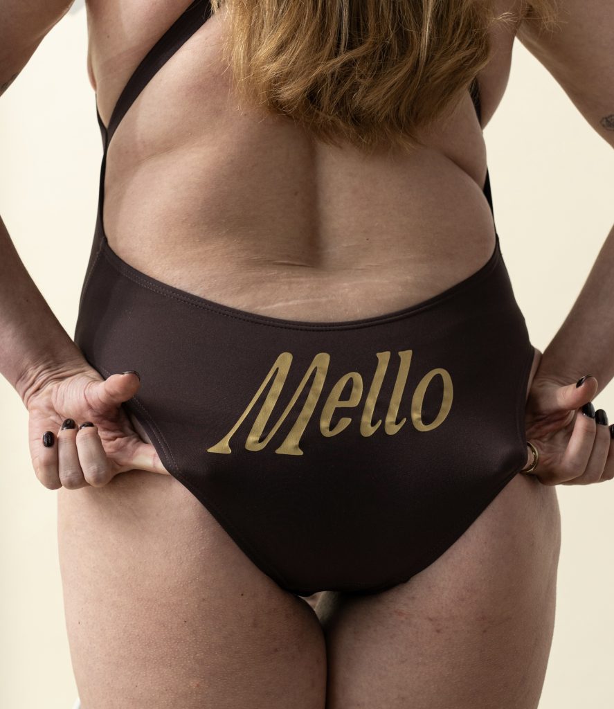 Back view of a woman wearing a brown Mello swimsuit with the Mello logo in gold across the back